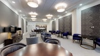 This thoughtfully designed community room offers a refined and welcoming environment suited for a variety of social occasions. The space is anchored by a drop-tile ceiling fitted with stylish drum pendant light fixtures and recessed lighting, providing warm, even illumination throughout. White-painted walls are accented by bold, decorative damask-patterned wall panels that add a sophisticated touch to the overall aesthetic.

The seating area is elegantly arranged with navy blue sofas and accent chairs alongside neutral gray upholstered chairs, centered around low round tables ideal for conversation and casual gatherings. Additional seating vignettes with armchairs and bistro-style tables line the walls, offering more intimate settings within the larger space.

At the far end of the room, a fully equipped kitchenette is visible, featuring dark cabinetry, modern appliances, and counter seating — making the space well-suited for catered events or light refreshments. A dining or conference table in rich wood tones is positioned in the foreground, further enhancing the room's versatility for both social and functional uses. Light-toned carpeting ties the design together, contributing to the space's polished, professional ambiance.