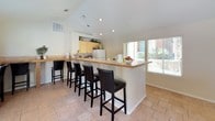 This single-story clubhouse offers a welcoming and functional space designed for community use. The open floor plan connects several key areas seamlessly, creating an environment well-suited for group gatherings and events. Vaulted ceilings rise throughout the main area, adding a sense of openness and volume to the space. Light tile flooring runs consistently across the entire floor, contributing to a clean, cohesive aesthetic.

The dining area features multiple high-top tables with bar-height seating, accommodating groups of varying sizes. A tile-surround fireplace anchors one end of the room, providing a warm focal point, and decorative shelving above displays greenery and accent pieces. Large windows fitted with blinds allow natural light to fill the space while maintaining privacy.

The kitchen area is equipped with light wood cabinetry, tile countertops, a standard range, refrigerator, and sink. An extended bar counter with bar-stool seating connects the kitchen to the main gathering area, making it practical for serving during events. Recessed lighting throughout supplements the natural light. The overall ambiance is casual, comfortable, and functional, making this clubhouse a versatile venue for community activities.