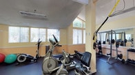 The fitness center at 9505 Gold Coast in San Diego is a well-appointed exercise facility designed to support a range of workout routines. The space features warm yellow and terracotta-toned walls that contribute to an energizing atmosphere, complemented by dark rubberized flooring ideal for exercise and equipment stability. Large windows along multiple walls flood the room with abundant natural light, offering views of surrounding greenery and creating an open, airy feel. The vaulted ceiling adds a sense of spaciousness to the facility. Structural columns are integrated throughout the layout, with one serving as an anchor point for TRX suspension training straps, making efficient use of available space. The equipment selection includes elliptical trainers, a rowing machine, a stationary recumbent bike, and a cable weight machine with a rack, catering to both cardiovascular and strength training needs. Mirrors along the walls further enhance the sense of space while allowing users to monitor their form. The overall layout is thoughtfully organized, providing ample room to move between stations comfortably. This fitness center offers residents a convenient and functional on-site option for maintaining an active lifestyle.