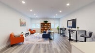 Located at 32 S Main St in downtown Concord, New Hampshire, this single-floor space offers a well-appointed layout comprising a living room and a dedicated office area. The interior features light gray luxury vinyl plank flooring throughout, crisp white walls, and recessed lighting that ensures consistent, bright illumination across the entire space.

The living room serves as the heart of the space, furnished with a navy blue sofa accented by vibrant throw pillows, bold orange armchairs, and a contemporary white coffee table. A sleek electric fireplace anchors one wall, complemented by colorful wall art above. A large wooden bookcase stocked with books, games, and decorative items sits between two generously sized windows that invite natural light and offer views of the surrounding Concord streetscape. High-top tables with modern bar stools provide additional seating along one wall, alongside a beverage station for added convenience.

The office area is practical and organized, featuring a dedicated desk setup and essential equipment. The entrance is defined by a glass-paneled gray door, a welcoming decorative wall sign, and a white reception counter, creating a professional yet inviting first impression. Together, the rooms create a cohesive, multi-functional environment ideal for both collaborative gatherings and focused work.