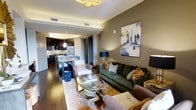 This single-floor residence showcases a thoughtfully designed open-concept layout where the living area and kitchen flow together seamlessly. The living room is anchored by a deep green sofa adorned with an eclectic mix of patterned throw pillows, complemented by a plush armchair and a round glass coffee table that rests atop a soft area rug. A striking abstract canvas in blue and gold tones decorates the taupe-painted accent wall, while a statement gold chandelier overhead adds a layer of glamour to the space. Recessed tray ceiling detailing adds architectural depth throughout.

Rich hardwood flooring runs continuously through the living space and into the kitchen, creating a cohesive aesthetic. The kitchen features dark cabinetry, a mosaic tile backsplash, stainless steel appliances, and a pendant-lit island that doubles as a dining area with seating for multiple guests. Navy blue accents — from the table lamp to decorative accessories — tie the living and kitchen areas together with a consistent color story. The overall ambiance is polished and inviting, blending contemporary elegance with warm, layered textures.