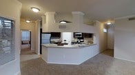 This single-story space showcases an open-concept layout where the kitchen serves as a central focal point. The kitchen is outfitted with dark espresso cabinetry that provides a striking contrast against the light-toned walls and countertops. A generous wraparound peninsula with a granite-style surface offers ample workspace and creates a natural division between the kitchen and the adjoining living area. Stainless steel appliances, including a refrigerator, electric range, and microwave, lend a modern, cohesive aesthetic to the cooking space. Decorative columns with crown molding detailing frame the kitchen area, adding an architectural dimension that elevates the overall design. A built-in range hood structure overhead adds a bold, angular visual element. The living area features plush wall-to-wall carpeting, contributing a sense of warmth and comfort. Flush-mount ceiling lighting provides consistent illumination throughout. The neutral color palette of whites and beiges across the walls and trim creates a clean, welcoming ambiance that ties the open floor plan together cohesively.