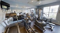 This fitness center offers a thoughtfully arranged workout space finished with light blue walls and rubber flooring that provides both durability and comfort underfoot. Large windows along one wall allow natural light to fill the room, creating a bright and energizing atmosphere for exercise. A ceiling fan overhead aids in air circulation, contributing to a comfortable environment during workouts.

The space is outfitted with a solid selection of cardio equipment, including elliptical trainers and a recumbent bike, catering to a range of aerobic exercise preferences. A multi-station cable machine is positioned toward the back of the room, supporting a variety of strength training exercises. A dedicated free weights area features a full rack of dumbbells in graduated sizes, ideal for targeted resistance training.

Full-length mirrors line one wall, visually expanding the space while allowing users to monitor their form during workouts. The overall layout is organized efficiently, ensuring ease of movement between equipment. The combination of natural light, practical flooring, and a diverse range of machines makes this fitness center a functional and well-rounded facility.