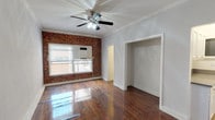 This single-floor space combines classic character with practical design throughout its thoughtfully arranged layout. The main living area is a standout, showcasing a striking exposed red brick accent wall that adds warmth and texture to the room. Richly toned hardwood floors run throughout, polished to a high sheen that reflects natural light pouring in from a large window. A ceiling fan with integrated lighting provides both comfort and illumination. An open archway leads to a secondary room, also finished with the same gleaming hardwood floors and its own ceiling fan, offering a versatile and well-lit space. Crown molding adds a refined architectural touch, while a built-in dresser with drawers provides ample storage. The bathroom is clean and classic, featuring full white subway tile surrounding a soaking tub with an overhead shower, accented by a decorative mosaic tile border. A window in the bathroom allows for natural light and ventilation. The kitchen is accessible from the main living area and features tile flooring. Throughout the space, white trim and neutral wall tones create a cohesive, bright atmosphere that highlights the character-rich architectural details, making this a distinctive and inviting place to call home.
