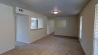 This single-story space presents an open and functional layout centered around a spacious carpeted living area. The main living space is finished with neutral-toned carpet and light-colored walls, creating a clean and understated atmosphere. A flush-mount ceiling light fixture provides overhead illumination, contributing to the room's practical design.

A notable architectural feature is the pass-through window connecting the living area to the kitchen, offering a glimpse into the brightly lit kitchen space beyond. The kitchen appears to feature white cabinetry and appliances, with the pass-through counter providing a convenient serving or casual dining surface.

The living area opens toward an additional room or hallway visible through a wide doorway opening on the far wall, suggesting a connected and accessible floor plan. A secondary window cutout or niche is visible on the adjacent wall, adding visual interest to the otherwise straightforward layout.

The overall ambiance is simple and neutral, offering a blank canvas adaptable to a variety of furnishing styles. The consistent use of light tones throughout the walls, ceiling, and flooring gives the space a cohesive and airy feel despite its modest scale.