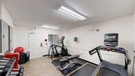 Located at 1920 Ivy Creek Blvd in Durham, NC, this fitness room offers a practical and well-equipped space for exercise. The room features light wood-look flooring throughout, white walls, and a textured ceiling illuminated by overhead fluorescent lighting, creating a clean and functional environment.

The cardio section includes a treadmill and an elliptical trainer, each positioned on protective rubber mats. A weight bench provides space for strength training, complemented by a three-tier dumbbell rack stocked with a range of free weights. A red mini-refrigerator offers convenient hydration access, while a set of red storage lockers along the back wall provides secure storage for personal belongings.

Natural light filters in through a window fitted with horizontal blinds, and a keyed exterior door offers a dedicated entry point to the room. Wall-mounted electrical panels are neatly housed on one wall, and safety features including a fire alarm panel and exit signage are present throughout. A wall-mounted media unit adds an entertainment option during workouts.

Overall, the space is efficiently arranged to maximize utility within its compact footprint, providing users with essential cardio and strength training equipment in a tidy, well-maintained setting.