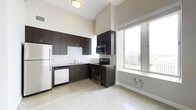 This single-floor space showcases a contemporary design with clean lines and a functional, open layout. The kitchen is a standout feature, outfitted with sleek dark espresso cabinetry that provides ample storage, complemented by light granite-style countertops and a classic white subway tile backsplash. Stainless steel appliances — including a refrigerator, electric range with microwave, and dishwasher — lend a polished, modern feel to the cooking area. A stainless steel sink sits beneath a window, adding convenience and natural light to the workspace. The flooring throughout is a speckled terrazzo-style surface, adding a subtle texture that brightens the overall interior. Soaring ceilings amplify the sense of openness, while a large multi-pane window along one wall floods the space with natural light and frames sweeping views of the surrounding cityscape. The neutral, light-toned walls enhance the airy atmosphere, creating a bright and welcoming environment. The overall ambiance is modern and urban, combining practical design elements with thoughtful architectural details that make the most of the available space and its elevated city setting.