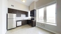 This single-floor space showcases a contemporary design with clean lines and a functional, open layout. The kitchen is a standout feature, outfitted with sleek dark espresso cabinetry that provides ample storage, complemented by light granite-style countertops and a classic white subway tile backsplash. Stainless steel appliances — including a refrigerator, electric range with microwave, and dishwasher — lend a polished, modern feel to the cooking area. A stainless steel sink sits beneath a window, adding convenience and natural light to the workspace. The flooring throughout is a speckled terrazzo-style surface, adding a subtle texture that brightens the overall interior. Soaring ceilings amplify the sense of openness, while a large multi-pane window along one wall floods the space with natural light and frames sweeping views of the surrounding cityscape. The neutral, light-toned walls enhance the airy atmosphere, creating a bright and welcoming environment. The overall ambiance is modern and urban, combining practical design elements with thoughtful architectural details that make the most of the available space and its elevated city setting.