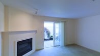 This single-level space presents a clean, understated living area designed for comfort and functionality. The room is finished with neutral, cream-toned walls and smooth ceilings that lend a bright, airy feel throughout. Soft wall-to-wall carpeting in a light beige hue covers the floor, adding warmth underfoot and contributing to the overall tranquil atmosphere.

A built-in fireplace with a sleek, modern surround serves as a focal point along one wall, providing both a practical heating source and a visually grounding element within the room. Track lighting mounted on the ceiling offers directional illumination to complement the abundant natural light that fills the space.

Sliding glass doors dominate the exterior wall, fitted with sheer vertical blinds that allow light to filter in softly while maintaining privacy. These doors open directly to a patio area, where mature ornamental trees and landscaping are visible, creating a seamless connection between the interior and the outdoors. The overall ambiance is calm and neutral, offering a versatile canvas well-suited to a variety of interior styles and furnishing choices.
