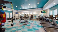 Located at 13444 Gran Bay Pkwy in Jacksonville, FL, this well-appointed fitness center is designed to deliver a comprehensive workout experience in a visually dynamic environment. The space is defined by its bold teal-painted ceiling and accent walls, which contrast sharply against crisp white walls and trim, creating an energetic yet polished atmosphere. Underfoot, a vibrant geometric-patterned floor in teal, gray, and white tones ties the entire space together with a modern aesthetic.

The cardio zone is generously equipped with multiple Cybex and True treadmills, elliptical machines, and stationary bikes, all arranged to allow ample personal space during use. A dedicated rowing machine and additional cable-based strength equipment occupy a separate alcove, complemented by motivational fitness artwork and a wall-mounted television. The strength training area features a multi-station cable machine, a full dumbbell rack spanning a wide weight range, and an adjustable weight bench, catering to users of all fitness levels.

Full-length mirrors line key walls throughout the space, amplifying the sense of openness and allowing users to monitor their form. Recessed lighting and multiple ceiling-mounted flat-screen televisions enhance the environment, while rows of windows fitted with blinds allow natural light to filter in. The single-floor layout ensures easy navigation across all workout zones.