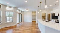 This thoughtfully designed single-floor residence offers a seamless blend of modern aesthetics and functional living. The open-concept kitchen and living room serve as the heart of the home, featuring warm hardwood-style flooring that extends throughout the main areas. The kitchen is appointed with crisp white shaker cabinetry with gold hardware, a curved quartz-topped island with an undermount sink, stainless steel appliances, and elegant brass pendant lighting that adds a refined touch. Built-in shelving provides convenient additional storage within the living area. Expansive windows with transom panels above and a sliding glass door flood the living space with natural light and offer outdoor views, enhancing the open, airy ambiance. The bedroom is carpeted for comfort, features a ceiling fan, and receives ample natural light through a large window. A spacious walk-in closet with multi-tier hanging rods and carpet flooring offers generous storage. Both bathrooms are finished with marble-style quartz countertops, white cabinetry, gold hardware and fixtures, and clean tile flooring. The unit also includes a dedicated laundry area, completing this well-appointed and move-in-ready residence.