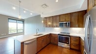 Located at 10730 Euclid Ave in Cleveland's University Circle neighborhood, this well-appointed one-bedroom, one-bathroom unit offers a thoughtfully designed single-floor layout. Rich hardwood flooring flows throughout the open-concept kitchen and living area, creating a warm and cohesive aesthetic. The kitchen is a standout feature, equipped with stainless steel appliances including a refrigerator, range, and microwave, complemented by rich wood cabinetry, a tile backsplash, white quartz countertops, and pendant lighting above the breakfast bar. The living area is bathed in natural light through floor-to-ceiling windows fitted with roller shades, and a glass door opens directly onto a private balcony with open urban views. The bedroom is carpeted for comfort and features a large window overlooking greenery, offering a tranquil retreat. A dedicated closet with louvered doors provides ample storage. The bathroom is finished with warm-toned tile flooring, a wood vanity with a white vessel sink, and a large mirror. Neutral taupe walls throughout create a calm, modern ambiance that ties each room together cohesively.
