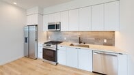 Located at 1112 Locust Ave in Long Beach, California, this modern single-floor residence showcases a clean, contemporary design throughout. Light oak hardwood flooring runs seamlessly across the entire space, complementing the crisp white walls and creating a bright, cohesive aesthetic.

The open-concept living and kitchen area is a standout feature, offering a generous, airy layout. The kitchen is outfitted with sleek flat-front white cabinetry extending to the ceiling, a neutral stone countertop, a subway-tiled backsplash in warm gray tones, and a full suite of stainless steel appliances including a gas range, built-in microwave, dishwasher, and refrigerator.

The living area flows naturally toward expansive black-framed corner windows and a glass sliding door, flooding the space with natural light and offering open views of the Long Beach skyline. Additional bedrooms continue the minimalist aesthetic, with large corner windows framing cityscape views and ample closet space, including louvered double-door closets with matte black hardware.

The bathrooms are elegantly finished with floor-to-ceiling white tile, glass-enclosed tub-shower combinations, matte black fixtures, and terrazzo-style flooring. Recessed lighting throughout enhances the modern, polished ambiance of this well-appointed Long Beach residence.