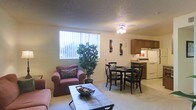 This single-floor residential space offers a thoughtfully arranged open-concept layout that seamlessly connects the living, dining, and kitchen areas into one cohesive and functional environment. The living area features plush seating, including a comfortable armchair, arranged around a tiled-top coffee table, creating a relaxed and welcoming atmosphere. A large window dressed with vertical blinds allows an abundance of natural light to fill the space, complemented by a ceiling-mounted light fixture overhead. A lush indoor plant adds a refreshing touch of greenery to the room. Adjacent to the living area, the dining space accommodates a round table with cross-back chairs, making it ideal for everyday meals or casual gatherings. The kitchen flows directly from the dining area and is equipped with wood-toned upper cabinetry, a white range with oven, a refrigerator, and ample counter space for meal preparation. The flooring throughout features neutral-toned carpet that adds warmth and continuity across the living and dining areas. The textured ceiling adds a classic architectural detail to the space. Overall, the unit presents a practical and comfortable layout suited for everyday living.