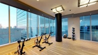 This Yoga & Spin Studio is a well-appointed fitness space designed to support a variety of workout disciplines in a polished, contemporary environment. The studio is defined by expansive floor-to-ceiling windows that wrap around the space, allowing abundant natural light to flood the interior and offering open views of the surrounding cityscape — creating an energizing atmosphere for exercise.

The flooring consists of light-toned hardwood, lending warmth and a clean aesthetic while providing a practical surface suitable for both spin and yoga activities. A row of professional spin bikes is positioned to take full advantage of the panoramic window views, making cycling sessions both motivating and visually engaging.

A bold, cylindrical structural column finished in matte black serves as a striking architectural focal point within the space. Overhead, a modern flush-mount light fixture provides supplemental ambient lighting alongside recessed ceiling lights, ensuring consistent illumination throughout.

Along the perimeter, a rack of medicine balls and a stability ball are neatly organized, offering functional accessories for strength and balance training. The overall design balances a contemporary aesthetic with practical fitness functionality, making it a versatile and inviting space for various wellness activities.