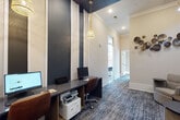 This single-floor business center is thoughtfully designed to balance productivity and comfort through a cohesive blend of contemporary and industrial aesthetics. The main workspace area features a built-in granite countertop workstation accommodating multiple computer terminals and a shared printer, set beneath soaring ceilings with bold navy accent panels and warm pendant lighting that add visual interest. Patterned blue-grey carpeting runs throughout, anchoring the space with texture and continuity. A standout private nook offers an individual granite desk positioned against a dramatic industrial-style mural wallcovering, complemented by a brass wall sconce and large windows that invite natural light and outdoor views — ideal for focused, independent work. Adjacent to the main area, a dedicated conference room provides a wood-grain table surrounded by upholstered navy chairs, a wall-mounted display screen, and a refined linear chandelier, making it well-suited for collaborative meetings or presentations. The lounge area features plush neutral armchairs paired with side tables, framed by large-scale abstract artwork and decorative metallic wall sculptures, creating an inviting space for informal gatherings. Together, the areas form a well-appointed, design-forward environment built for both individual focus and group collaboration.