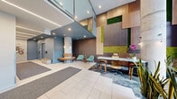 This contemporary building lobby makes a strong first impression with its soaring double-height ceilings and expansive floor-to-ceiling glass entryway, flooding the space with natural light and offering clear views to the streetscape beyond. The entry vestibule is framed by exposed brick walls and large-format tile flooring, transitioning seamlessly into the main lobby area. A standout design feature is the striking accent wall composed of warm wood panels interspersed with vibrant living moss installations, adding a dynamic, biophilic element to the interior. The lobby is thoughtfully zoned into distinct functional areas: a casual lounge seating area centered around a circular bench and a pair of sage green armchairs, and a co-working station featuring a sleek marble-topped counter with ergonomic bar-height seating. Recessed and linear LED lighting throughout creates a clean, polished ambiance. Decorative pendant lights and fresh orchids add refined finishing touches. A corridor leading to the elevators is adorned with a large, colorful abstract artwork, contributing to the lobby's curated aesthetic. The combination of natural materials, greenery, and modern furnishings creates a welcoming, professional environment that balances functionality with thoughtful design.