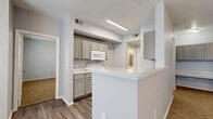 This single-story, one-bedroom, one-bathroom residence offers an efficient and functional layout across a well-utilized footprint. The interior is finished with a neutral gray color palette throughout, creating a cohesive and understated aesthetic.

The kitchen is positioned centrally within the unit and features gray shaker-style cabinetry, a white appliance suite including a range and over-the-range microwave, and a light-toned countertop that extends to form a peninsula breakfast bar. The peninsula separates the kitchen from the adjacent living area, providing both a visual boundary and additional workspace. Wood-look plank flooring runs through the kitchen and hallways, adding durability and a modern touch.

The living area is generously proportioned for the overall unit size, featuring carpet flooring, a ceiling fan with integrated light fixture, and multiple windows fitted with horizontal blinds that allow for ample natural light. A hallway connects the main living space to the bedroom and bathroom.

The bedroom offers comfortable dimensions, with carpet flooring, a ceiling fan, a closet, and a window providing natural light. The full bathroom includes a single-sink vanity with gray cabinetry matching the kitchen, a lighted mirror, and a full tub-shower combination with white tile surround and wood-look flooring.