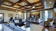 This single-floor space makes a striking first impression with its beautifully crafted coffered ceiling, adorned with rich dark wood beams that add depth, warmth, and architectural character to the interior. Pendant lantern-style light fixtures hang gracefully from the ceiling, complementing the recessed lighting throughout and casting a warm, inviting glow across the open floor plan.

The expansive living area is anchored by plush, dark upholstered sofas arranged around a classic pedestal coffee table, all set atop a neutral area rug over dark hardwood flooring. Soft decorative pillows and layered seating create a comfortable and welcoming atmosphere.

Seamlessly connected to the living space is a well-appointed kitchen featuring warm wood cabinetry, stainless steel appliances, and a generous wraparound breakfast bar with elegant wrought iron bar stools. The kitchen's layout is both functional and stylish, offering ample counter space for cooking and entertaining.

Natural light filters in through arched and French-style windows dressed with soft draping curtains, adding an airy quality that balances the richness of the dark wood accents. The overall ambiance is refined yet relaxed, blending traditional craftsmanship with comfortable, everyday livability throughout this thoughtfully designed space.