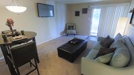 This single-floor space offers a welcoming and functional layout designed for comfortable everyday living. The living room serves as the central gathering area, featuring plush wall-to-wall carpeting that adds warmth underfoot. A wall-mounted flat-screen television anchors one side of the room, creating a natural focal point for the space. Seating is arranged thoughtfully around a tufted dark leather ottoman that doubles as a coffee table, accompanied by a light-colored sofa and a classic striped accent chair nestled in the corner, adding a touch of character to the room. A small bistro-style table and chair set near the edge of the space provides an intimate spot for casual dining or morning coffee. Natural light filters generously into the room through a sliding glass door fitted with vertical blinds, offering a view of the greenery outside and a seamless connection to the outdoors. The neutral-toned walls and clean baseboards give the space a tidy, well-maintained appearance. Subtle wall décor adds a modest artistic touch without overwhelming the understated aesthetic. Overall, the space presents a cozy, practical environment with a calm and neutral ambiance suited for relaxed, everyday living.