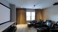 This single-floor space exudes a polished, upscale ambiance designed for comfort and relaxation. The main living or lounge area is anchored by a collection of dark leather armchairs with classic nail-head trim detailing, accompanied by matching ottomans that invite leisurely seating. The room's refined aesthetic is enhanced by sweeping floor-to-ceiling gold-toned drapes that frame large windows, allowing natural light to filter softly into the space while maintaining an air of elegance. The light-toned hardwood flooring adds warmth and contrast against the neutral gray walls, creating a balanced and inviting palette. Overhead, a globe pendant light fixture serves as a subtle yet stylish focal point, complemented by recessed lighting that provides even, ambient illumination throughout. A media console or entertainment unit is positioned along one wall, suggesting the space may function as a screening or media lounge. The overall design strikes a harmonious balance between classic and contemporary elements, making it well-suited for both quiet relaxation and social gatherings. The thoughtful arrangement of furnishings and the layered lighting design contribute to a space that feels both functional and distinctly sophisticated.