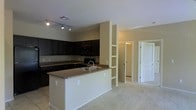 This single-story space showcases an open-concept layout that integrates the kitchen and living areas into one cohesive environment. The kitchen is a standout feature, outfitted with rich, dark espresso-toned cabinetry that contrasts sharply against the light-colored walls. Granite countertops run throughout, including along the peninsula-style island, which is equipped with a double-basin sink and provides additional workspace and casual seating potential. Black appliances complement the darker cabinetry, maintaining a consistent color palette. Track lighting on the ceiling ensures the kitchen is well-illuminated. A built-in shelving column serves as a subtle architectural divider between the kitchen and the adjacent living space, offering both display and storage functionality. The transition from tile flooring in the kitchen to plush carpeting in the living area defines each zone while preserving the open feel of the layout. Doorways visible from the kitchen suggest additional rooms beyond, indicating a practical and well-connected floor plan. The neutral wall tones and standard ceiling height contribute to a clean, functional ambiance throughout.