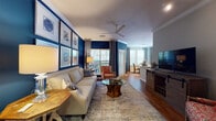Located at 1100 Legends Club Dr in Mount Pleasant, SC, this single-floor residence offers a well-appointed layout designed for comfortable living. The living room is a standout space, featuring rich hardwood floors, crown molding, and a bold navy blue accent wall adorned with a gallery of framed botanical artwork. A ceiling fan, ample natural light from large windows, and a distinctive glass-top coffee table with a natural wood base anchor the room's coastal-inspired aesthetic. The space flows seamlessly into a dedicated dining area, also highlighted by the striking navy accent wall and natural light. The kitchen is outfitted with stainless steel appliances, white cabinetry, stone-look countertops, a subway tile backsplash, and a breakfast bar peninsula. The primary bedroom offers plush carpeting, a decorative chandelier, sliding glass door access to an outdoor area, and an ensuite bathroom with a walk-in closet. A second full bathroom features hardwood flooring, a tub-shower combination, and convenient closet access. A dedicated laundry room with wire shelving provides functional storage. The property also includes access to an elegantly designed community lounge with soaring ceilings, a coffered ceiling detail, barn doors, and floor-to-ceiling windows.