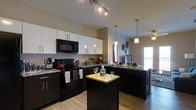 This single-floor space showcases a thoughtfully designed, open-concept kitchen that serves as the focal point of the home. The kitchen features a striking contrast of dark lower cabinetry paired with crisp white upper cabinets, all complemented by granite countertops with subtle veining. Black appliances, including a range, built-in microwave, and refrigerator, maintain a cohesive, modern aesthetic throughout the space.

A freestanding kitchen island with a butcher block top provides additional workspace and casual seating, while an extended peninsula with a sink and dark cabinetry offers even more functional counter space. Overhead, a combination of track lighting and elegant pendant lights illuminate both the kitchen and the adjoining areas, creating a warm and inviting atmosphere.

The light hardwood flooring adds warmth and continuity, flowing seamlessly from the kitchen toward the adjacent living areas. A glimpse of a patio door in the background suggests access to an outdoor space, allowing natural light to filter into the open layout. The overall design balances functionality with a contemporary, polished style.