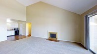This single-floor space makes a strong first impression with its soaring vaulted ceiling, which dramatically increases the sense of volume in the main living area. The steep pitch of the ceiling draws the eye upward, creating an expansive, open feel that distinguishes the space from standard flat-ceiling designs. Neutral beige walls complement the light-toned carpet underfoot, contributing to a warm and understated aesthetic throughout.

A freestanding fireplace with a gold-toned surround and brick hearth serves as a natural focal point along one wall, adding a classic touch and offering a functional source of warmth. The fireplace's traditional detailing provides visual interest against the otherwise clean, minimalist wall surfaces.

A glimpse into an adjacent hallway or room reveals hardwood-style flooring and a warm-toned door, suggesting a transition into additional areas of the home. The layout appears open and versatile, well-suited for a variety of furniture arrangements. Overall, the space combines architectural character—particularly through its vaulted ceiling—with simple, neutral finishes that offer a blank canvas for personalization.