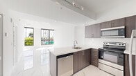 Located at Hook Square in Miami Springs, FL, this well-appointed two-bedroom, two-bathroom unit offers a thoughtfully designed open-concept layout across a single floor. The interior is defined by large-format polished porcelain tiles with a marble-like veining pattern that runs consistently throughout, reflecting natural light and lending a clean, contemporary aesthetic to every room.

The open-concept living and kitchen area serves as the heart of the unit. The kitchen features dark wood-grain cabinetry, a white quartz peninsula with an undermount sink, and a full suite of stainless steel appliances including a dishwasher, range, microwave, and refrigerator. Track lighting overhead provides functional illumination, while the open connection to the living area creates an airy, sociable atmosphere.

Both bedrooms are generously proportioned, with the spacious primary bedroom offering ample room for furnishings. Floor-to-ceiling windows in the bedrooms flood the spaces with natural light and frame views of the surrounding greenery and Miami Springs streetscape. Closet storage is provided with wire-shelf systems.

The bathrooms are finished with marble-look tile, including a walk-in shower with full-height stone-style tile surround and chrome fixtures, maintaining the cohesive modern aesthetic found throughout the unit.