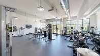 Located at 409 East Butler Avenue in New Britain, PA, this fitness center offers a thoughtfully designed, single-floor exercise space that accommodates a comprehensive range of workout needs. The facility features soaring ceilings with exposed industrial-style ductwork and stylish pendant lighting, lending the space a modern, open atmosphere. Large floor-to-ceiling windows along the perimeter flood the interior with abundant natural light and provide views of the surrounding greenery, creating an energizing environment for exercise.

The main workout area is fitted with durable rubber flooring and houses a full lineup of cardio equipment, including multiple treadmills and upright and recumbent exercise bikes. Strength training is well supported by a multi-station cable and weight machine, a dedicated dumbbell rack with a wide weight range, and wall-mounted mirrors to assist with form. Wall-mounted flat-screen televisions are positioned for convenient viewing during workouts.

A distinct functional training zone transitions to hardwood-style flooring and is equipped with a cable functional trainer, stability and medicine balls on a dedicated rack, foam rollers, and a plyo box — ideal for dynamic and mobility-focused training. The overall layout is spacious and uncluttered, allowing users to move freely between equipment zones while enjoying a clean, well-maintained facility.