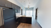 This contemporary single-floor unit offers a well-designed open-concept layout that seamlessly connects the kitchen, dining, and living areas. The kitchen is outfitted with dark espresso cabinetry, light countertops, and a full suite of black appliances including a refrigerator, electric range, microwave, and dishwasher. A peninsula with an integrated sink serves as both a functional workspace and a natural divider between the kitchen and living area. Stylish pendant lighting adds a modern touch overhead, while a ceiling fan keeps the living space comfortable. Large sliding glass doors open to a private balcony, allowing abundant natural light to fill the open area. Wood-style flooring runs consistently throughout the entire unit, complemented by dark baseboards that create a clean, modern contrast against the light-toned walls. The two bedrooms are well-proportioned, each featuring a window for natural light and a ceiling fixture. Both bathrooms are thoughtfully appointed with dark vanity cabinetry, light countertops, and a bathtub, and each connects directly to a dedicated walk-in closet with built-in shelving and hanging rods, providing excellent storage and organization throughout the unit.