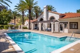 Located at Sea Cliff Way in Oceanside, California, this resort-style outdoor amenity space offers a compelling combination of recreational facilities and Southern California landscaping. The centerpiece is a generously sized rectangular lap pool with sparkling turquoise water and tiled edges, designed for both fitness and leisure. Adjacent to the pool sits a separate spa with easy step-in access and dedicated lounge chairs and umbrellas for relaxing poolside.

The grounds are richly landscaped with towering mature palm trees and lush greenery that provide both shade and a tropical ambiance, all set against a backdrop of natural hillside terrain. A dedicated BBQ station with an outdoor grill and seating area extends the utility of the space for social gatherings. Multiple patio lounge chairs and shaded umbrella stations are thoughtfully distributed around the deck.

The Spanish Colonial Revival-style clubhouse building anchors the space architecturally, featuring a white stucco exterior, terracotta tile roofline, arched windows, a prominent chimney, and a pergola-covered entrance. The building opens directly onto the pool deck, integrating indoor and outdoor spaces seamlessly. Under the clear blue skies of Southern California, this amenity area delivers a vibrant, resort-like atmosphere year-round.