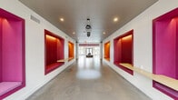 Located at 723 Wheatland St in Phoenixville, PA, this single-floor space makes a striking first impression with its bold, contemporary design. The expansive hallway serves as a dynamic spine of the layout, lined on both sides with large, deeply recessed wall niches upholstered in vivid red and orange hues, each equipped with built-in wooden shelves. Polished concrete floors run throughout, reflecting recessed ceiling lighting that keeps the space bright and welcoming. The entry foyer features floor-to-ceiling glass doors that flood the area with natural light and offer views of the outdoor surroundings, while bold magenta ceiling beams add a distinctive architectural accent. A dedicated office and meeting area is furnished with a conference table and ergonomic seating alongside a lounge zone, all anchored by vibrant teal carpeting. Large windows in this room overlook an outdoor pool area, enhancing the connection to the exterior. Walls feature curated artwork and typographic installations, reinforcing the space's communicative, community-oriented character. An interior gallery wall with pink-backed display niches and a bold yellow accent door contribute further pops of color. Throughout, the design balances energetic visual identity with functional, flexible areas suited for gathering, working, and socializing.