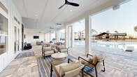 Located at 500 Liberty Ln NW in Fort Walton Beach, Florida, this single-level outdoor amenity space is designed with a coastal-inspired aesthetic that blends relaxation and recreation seamlessly. The centerpiece is a expansive resort-style swimming pool with a smooth, curved edge and a generous stone-tile surround, offering ample space for sunbathing on modern lounge chairs. A covered pavilion anchors one end of the pool deck, featuring exposed wood beam construction, a stone fireplace, and a built-in grill — ideal for outdoor gatherings. The covered clubhouse patio extends along the pool's edge, finished with large-format stone tile flooring and outfitted with ceiling fans throughout. Multiple seating arrangements with upholstered chairs and sofas invite relaxed conversation, while a dedicated game area includes foosball and a ping-pong table alongside a wall-mounted TV. Shiplap-style white paneling, floor-to-ceiling glass doors, and wide open archways define the architectural character of the covered areas, creating a bright and airy atmosphere. A spacious open-air patio extends beyond the covered zone, enclosed by ornamental black iron fencing with manicured landscaping. The surrounding residential buildings frame the space, contributing to a vibrant community atmosphere.