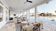 Located at 500 Liberty Ln NW in Fort Walton Beach, Florida, this single-level outdoor amenity space is designed with a coastal-inspired aesthetic that blends relaxation and recreation seamlessly. The centerpiece is a expansive resort-style swimming pool with a smooth, curved edge and a generous stone-tile surround, offering ample space for sunbathing on modern lounge chairs. A covered pavilion anchors one end of the pool deck, featuring exposed wood beam construction, a stone fireplace, and a built-in grill — ideal for outdoor gatherings. The covered clubhouse patio extends along the pool's edge, finished with large-format stone tile flooring and outfitted with ceiling fans throughout. Multiple seating arrangements with upholstered chairs and sofas invite relaxed conversation, while a dedicated game area includes foosball and a ping-pong table alongside a wall-mounted TV. Shiplap-style white paneling, floor-to-ceiling glass doors, and wide open archways define the architectural character of the covered areas, creating a bright and airy atmosphere. A spacious open-air patio extends beyond the covered zone, enclosed by ornamental black iron fencing with manicured landscaping. The surrounding residential buildings frame the space, contributing to a vibrant community atmosphere.