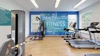 This fitness center is thoughtfully designed to support a comprehensive workout experience across two connected zones. The main exercise area features light hardwood-style flooring, bright white walls, and an abundance of natural light streaming through multiple large windows. Ceiling fans keep the space comfortable during intense workouts. A bold, eye-catching blue accent wall emblazoned with fitness-themed typography — featuring words like "Health," "Fitness," "Energy," and "Wellbeing" — adds a motivating and energetic atmosphere to the room. The main floor is equipped with multiple treadmills, elliptical cross-trainers, and a cable weight machine, providing ample options for both cardio and strength training. Recessed and flush-panel fluorescent lighting ensures the space remains well-lit at all times. A separate studio area, accessible from the main room, features vaulted ceilings with a skylight, a large floor-to-ceiling mirror wall, and space for flexibility and functional training. This area includes stability balls, medicine balls, resistance equipment, and a yoga mat, making it ideal for stretching or guided workouts. Large glass-paned doors open directly to an outdoor pool area, seamlessly blending indoor fitness with outdoor recreation.