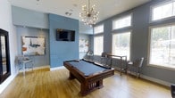 This contemporary recreation lounge is designed for both socializing and leisure, combining modern aesthetics with functional amenities. The space is anchored by a rich, dark-wood billiards table set atop warm hardwood flooring, serving as the room's central focal point. Overhead, a striking Sputnik-style chandelier adds a bold mid-century modern design element, casting a warm ambient glow throughout the room.

The walls are finished in a cool, calming blue-gray tone that lends the space a sophisticated and inviting atmosphere. A wall-mounted flat-screen television is positioned prominently on the feature wall, making the room well-suited for watching sports or entertainment during gatherings. Curated artwork adorns the walls, adding a gallery-like quality to the space.

Large windows along one wall flood the interior with abundant natural light and offer views to the outdoors, enhancing the open, airy feel of the room. A high-top bar table with modern counter-height stools is positioned near the windows, providing a casual seating area ideal for conversation or spectating. The elevated ceiling height further contributes to the spacious, generous feel of this well-appointed amenity space.