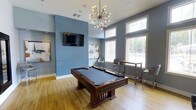 This contemporary recreation lounge is designed for both socializing and leisure, combining modern aesthetics with functional amenities. The space is anchored by a rich, dark-wood billiards table set atop warm hardwood flooring, serving as the room's central focal point. Overhead, a striking Sputnik-style chandelier adds a bold mid-century modern design element, casting a warm ambient glow throughout the room.

The walls are finished in a cool, calming blue-gray tone that lends the space a sophisticated and inviting atmosphere. A wall-mounted flat-screen television is positioned prominently on the feature wall, making the room well-suited for watching sports or entertainment during gatherings. Curated artwork adorns the walls, adding a gallery-like quality to the space.

Large windows along one wall flood the interior with abundant natural light and offer views to the outdoors, enhancing the open, airy feel of the room. A high-top bar table with modern counter-height stools is positioned near the windows, providing a casual seating area ideal for conversation or spectating. The elevated ceiling height further contributes to the spacious, generous feel of this well-appointed amenity space.