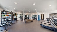 Located at 2058 East Apache Boulevard in Tempe, Arizona, this fitness center offers a generously sized, single-floor exercise space designed to support a wide variety of workout routines. The open floor plan is finished with durable speckled rubber flooring throughout, providing both safety and functionality for high-intensity training.

The strength training area is extensively equipped with cable machines, a multi-function rack system, barbells, and an organized dumbbell rack with a full range of free weights. A dedicated functional training zone features battle ropes, resistance bands, a punching bag, and a wall-mounted television, complemented by large windows that bring in natural light and outdoor views. Floor-to-ceiling mirrors line key walls, visually expanding the space and allowing users to monitor their form.

The cardio section houses multiple treadmills, a stair climber, a stationary bike, and a rowing machine, all positioned near expansive floor-to-ceiling windows that overlook the surrounding streetscape. A sturdy wooden bench sits centrally, serving as a versatile resting and workout station.

Additional amenities include a set of blue storage lockers, recessed ceiling lighting throughout, and a clean, modern white interior that creates a bright, motivating atmosphere. The space is thoughtfully organized to allow fluid movement between training zones.