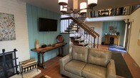 This two-story space blends rustic and southwestern design elements into a cohesive, character-rich interior. The main living area features teal-painted shiplap walls that lend a warm, cottage-like atmosphere, complemented by terracotta tile flooring that adds earthy texture underfoot. A wall-mounted flat-screen television is positioned above a dark wood console table with turned legs, providing both function and traditional style.

A statement wrought iron and wood staircase serves as a focal centerpiece, featuring ornate scrollwork balusters and open wood treads that connect the two levels with visual elegance. The upper level, visible from below, features white painted brick walls, adding architectural contrast and texture to the space.

Overhead, an iron chandelier with cylindrical shades provides ambient lighting that enhances the rustic aesthetic. The seating area is anchored by a light gray tufted leather loveseat, offering a neutral balance to the warm tones throughout. The overall ambiance is inviting and layered, combining traditional furnishings with Southwestern architectural details for a distinctive, well-appointed interior.