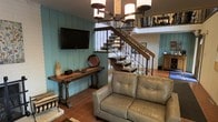 This two-story space blends rustic and southwestern design elements into a cohesive, character-rich interior. The main living area features teal-painted shiplap walls that lend a warm, cottage-like atmosphere, complemented by terracotta tile flooring that adds earthy texture underfoot. A wall-mounted flat-screen television is positioned above a dark wood console table with turned legs, providing both function and traditional style.

A statement wrought iron and wood staircase serves as a focal centerpiece, featuring ornate scrollwork balusters and open wood treads that connect the two levels with visual elegance. The upper level, visible from below, features white painted brick walls, adding architectural contrast and texture to the space.

Overhead, an iron chandelier with cylindrical shades provides ambient lighting that enhances the rustic aesthetic. The seating area is anchored by a light gray tufted leather loveseat, offering a neutral balance to the warm tones throughout. The overall ambiance is inviting and layered, combining traditional furnishings with Southwestern architectural details for a distinctive, well-appointed interior.
