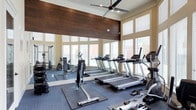 This fitness center is designed to offer a comprehensive workout experience within a bright, open environment. The space features soaring ceilings accented by crown molding and a large ceiling fan that promotes air circulation throughout. A striking wall of dark wood paneling serves as a bold architectural focal point, adding warmth and contrast to the otherwise light-toned interior.

Floor-to-ceiling windows line the walls, flooding the space with abundant natural light while offering views of the surrounding exterior. Slatted blinds allow for adjustable light control throughout the day. The flooring consists of durable, commercial-grade carpet tiles in a charcoal blue tone, with rubberized fitness mats positioned beneath equipment for added safety and floor protection.

The cardio area is well-appointed with a row of modern treadmills and elliptical machines positioned to face the windows, providing an engaging view during workouts. A dedicated strength training zone includes a cable machine, weight bench, and a dumbbell rack. Wall-mounted mirrors expand the perceived scale of the space while allowing users to monitor their form. Overall, the fitness center presents a clean, functional, and inviting atmosphere.