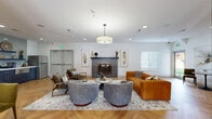 This single-floor amenity lounge is designed to serve as a versatile communal gathering space, blending comfort and functionality through thoughtful design. The entire area is unified by striking herringbone-patterned hardwood flooring, which runs continuously throughout every zone and contributes warmth and visual cohesion to the open layout.

The central lounge area serves as the focal point of the space, anchored by a textured tile fireplace framed with decorative artwork. A mix of seating — including rounded swivel chairs in heathered grey, a cognac leather sofa, and mid-century accent chairs — surrounds a marble-topped coffee table, creating an inviting conversation area beneath a large drum pendant light.

Adjacent to the lounge, a casual dining and seating zone features bistro-style round tables paired with upholstered chairs, illuminated by matte black pendant lights. Large windows throughout admit ample natural light and offer views of the surrounding greenery.

A well-appointed wet bar station includes charcoal shaker-style cabinetry with brushed gold hardware, a white quartz countertop, undermount sink, stainless steel appliances, and open shelving backed by a light grey subway tile. A full-size stainless steel refrigerator is also available nearby.

A dedicated game area houses a foosball table, completing this multi-functional space designed for both relaxation and recreation.