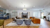 This single-floor amenity lounge is designed to serve as a versatile communal gathering space, blending comfort and functionality through thoughtful design. The entire area is unified by striking herringbone-patterned hardwood flooring, which runs continuously throughout every zone and contributes warmth and visual cohesion to the open layout.

The central lounge area serves as the focal point of the space, anchored by a textured tile fireplace framed with decorative artwork. A mix of seating — including rounded swivel chairs in heathered grey, a cognac leather sofa, and mid-century accent chairs — surrounds a marble-topped coffee table, creating an inviting conversation area beneath a large drum pendant light.

Adjacent to the lounge, a casual dining and seating zone features bistro-style round tables paired with upholstered chairs, illuminated by matte black pendant lights. Large windows throughout admit ample natural light and offer views of the surrounding greenery.

A well-appointed wet bar station includes charcoal shaker-style cabinetry with brushed gold hardware, a white quartz countertop, undermount sink, stainless steel appliances, and open shelving backed by a light grey subway tile. A full-size stainless steel refrigerator is also available nearby.

A dedicated game area houses a foosball table, completing this multi-functional space designed for both relaxation and recreation.