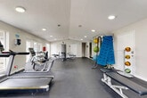 This fitness center offers a comprehensive workout environment within a single, open-plan floor. The space is finished with durable dark rubber flooring throughout, providing a practical and professional surface suited to a wide range of exercise activities. Recessed lighting is distributed evenly across the textured ceiling, ensuring the entire area is well-lit, while multiple windows and glass-paneled doors along the perimeter admit generous natural light and offer views of the outdoor pool area beyond.

The cardio zone features multiple treadmills and a stationary cycle, arranged to take advantage of natural light from the exterior-facing windows. A full-length wall mirror runs along one side of the room, visually expanding the space and aiding proper form during workouts. The strength training area includes a multi-station cable machine, a flat utility bench, and a dumbbell rack stocked with a range of weights.

A dedicated functional training station houses an array of stability balls, battle ropes, resistance bands, and a yoga/exercise mat, supporting dynamic and flexibility-based training routines. The clean white walls and open floor plan create an uncluttered, motivating atmosphere. The layout allows for comfortable movement between equipment zones, making this fitness center a versatile and well-appointed amenity.