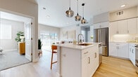 This single-floor residence showcases a thoughtfully designed open-concept layout that seamlessly connects the kitchen, dining, and living areas. The kitchen is a standout feature, equipped with full-height white shaker cabinetry, quartz countertops, and a suite of stainless steel appliances including a French door refrigerator, electric range, and built-in microwave. A generously sized central island with an undermount sink and pendant lighting serves as both a functional workspace and a natural gathering point. Light wood-tone flooring flows throughout the main living areas, enhancing the bright, airy atmosphere. The living room opens directly to an outdoor balcony through sliding glass doors with vertical blinds, allowing abundant natural light to fill the space. Two bedrooms feature plush carpeting and sliding closet doors, offering comfortable retreats. The unit includes two bathrooms: one with a soaking tub and shower combination, and another with a standalone glass-enclosed shower. Both bathrooms feature wood-look flooring, white vanities with quartz counters, and built-in shelving for storage. The overall design aesthetic is clean, modern, and cohesive throughout.