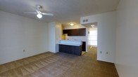 This single-floor space offers an open-concept layout that seamlessly connects the living area to the kitchen. The living room is finished with wall-to-wall carpeting in a neutral tan tone, creating a warm and comfortable foundation throughout the space. A ceiling fan with integrated light fixture is centrally mounted on the textured ceiling, providing both air circulation and ambient lighting.

The kitchen is defined by dark espresso-toned cabinetry that contrasts with light countertops, offering a modern aesthetic. A breakfast bar extends from the kitchen counter, providing a casual dining or workspace option that further reinforces the open flow between the kitchen and living areas. A range hood is visible beneath the upper cabinets, indicating a dedicated cooking space.

A hallway visible beyond the kitchen leads to additional areas of the unit, with a window at the far end allowing natural light to filter through. Overhead lighting and a ceiling vent ensure functional climate control throughout. The textured ceiling and neutral wall tones contribute to a clean, understated interior aesthetic overall.
