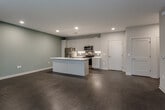 This single-floor unit in North Kansas City, Missouri, presents a contemporary design with a thoughtful, functional layout. The open-concept living and kitchen area serves as the centerpiece of the space, featuring dark hardwood-style flooring, recessed lighting, and a neutral color palette with soft blue-gray accent walls. A ceiling fan adds comfort to the spacious living area, which receives natural light through large windows fitted with horizontal blinds.

The kitchen is well-appointed with white cabinetry reaching toward the ceiling, a white countertop island with a sink, stainless steel appliances including a refrigerator, electric range, and over-the-range microwave, and a subway tile backsplash. An in-unit stacked washer and dryer is conveniently accessible off the main living area.

The additional rooms feature plush carpeting or hardwood-style flooring, neutral toned walls, and windows that allow in natural light. The bathroom is finished in a clean, modern style with large-format tile flooring, a sliding glass-door shower, a white elongated vanity with ample storage, and a mirrored medicine cabinet. Overall, the space offers a cohesive, move-in-ready aesthetic with modern finishes throughout.