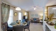 This single-story residence features an open-concept layout that seamlessly connects the dining and living areas, creating a cohesive and welcoming environment. The dining area is anchored by a round glass-top table surrounded by upholstered chairs with nail-head trim detailing, positioned beneath a dome pendant light that adds a focused, warm glow. Natural light filters in through a nearby door with blinds and patterned curtains, enhancing the bright, airy feel of the space. A decorative hexagonal mirror adorns the wall, adding visual interest. The living area flows naturally from the dining space, centered around a plush sofa accented with teal and patterned throw pillows. A ceiling fan with an integrated light fixture keeps the space comfortable, while soft carpet underfoot adds warmth and texture. A media console along one wall houses a flat-screen television, complemented by ambient floor and table lamps that contribute to the room's inviting atmosphere. Neutral wall tones throughout provide a versatile backdrop, while teal and earthy accent colors in the furnishings and artwork tie the two areas together cohesively.